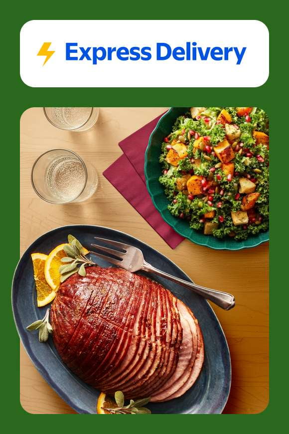 Express Delivery. A platter with a sliced ham and orange slices and a bowl of kale and roasted root vegetables on a wood countertop.