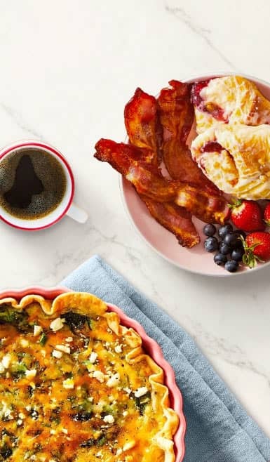 A quiche is shown next to a plate of bacon, croissant, fruit and a cup of coffee.