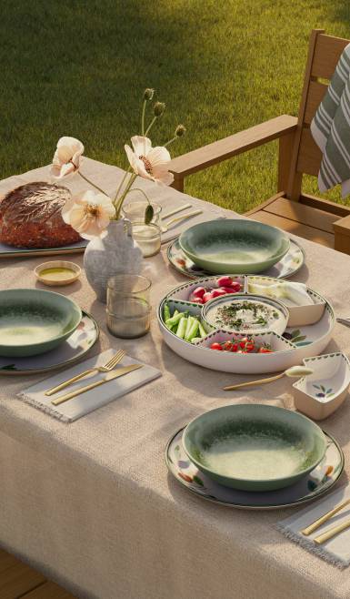 Outdoor table set with green bowls, floral plates, bread, vegetables, dip platter, glasses, and flowers in soft evening sunlight.