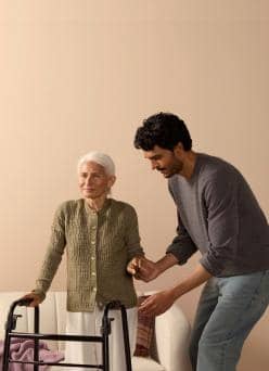 An older adult uses a walker beside a couch while a younger adult stands close, holding their hand for support.
