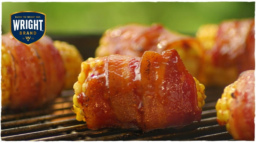 Mini corn on the cob wrapped in sizzling Wright Brand Bacon while roasting on a grill top.