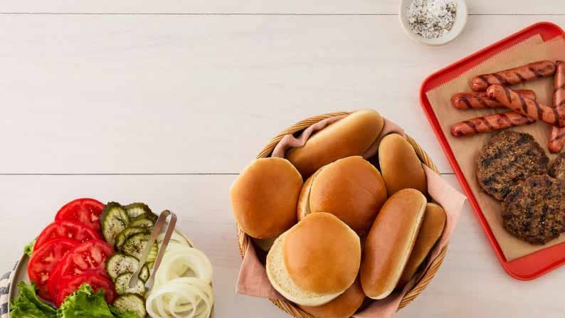 A plate with sliced vegetables, a plate with hamburger and hot dog buns, and a red tray with grilled hamburger patties and hot dogs.