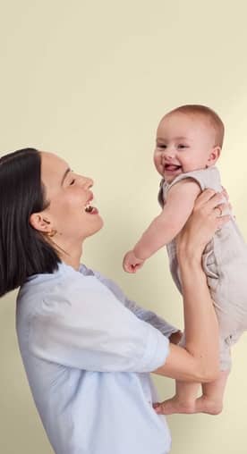 Mom holding her baby smiling in front of a crib. The Baby Event badge in pink.