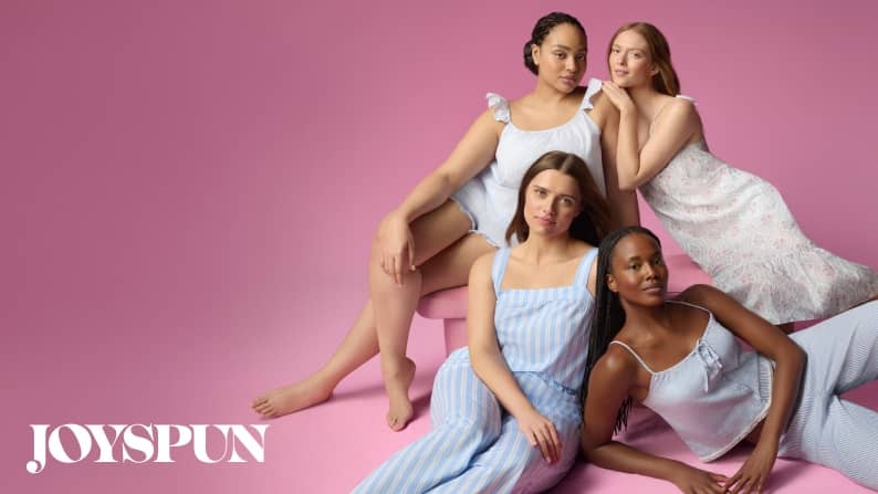 From Joyspun: four women in pastel pajamas sit together against a soft pink backdrop, smiling and showcasing diverse body types and cozy pajamas.