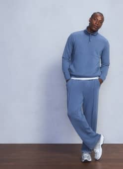 Man leaning against a wall wearing a blue quarter-zip pullover, matching track pants and white sneakers.