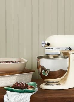 A cream-colored stand mixer is on a wood countertop next to a beige bakingdish and a green plate with holiday cookies.