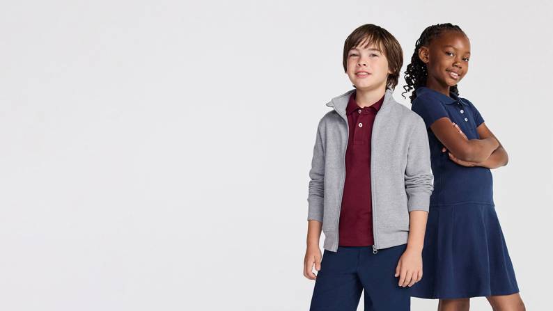 A boy in a burgundy polo and navy trousers next to a girl in a short-sleeved navy polo uniform dress.
