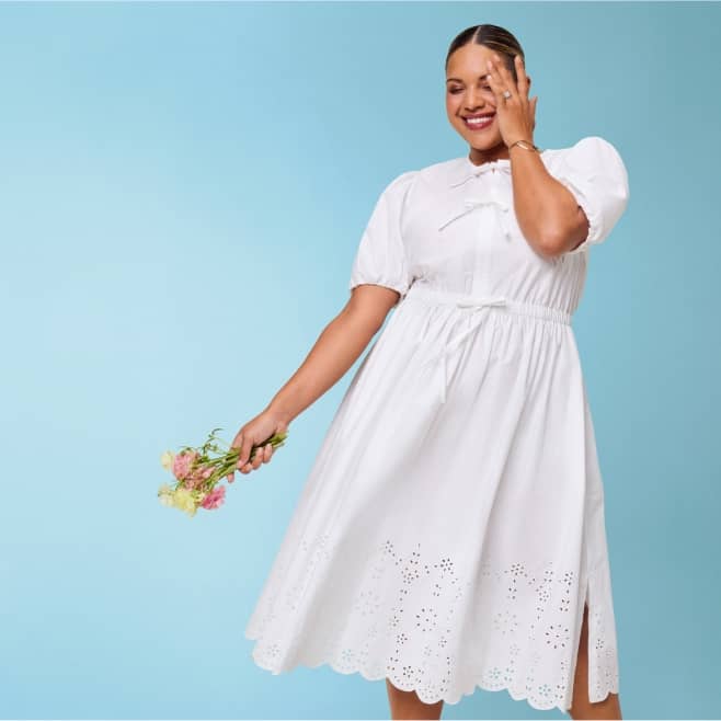 A woman wearing a white dress and holding flowers is pictured standing in front of a blue backdrop.