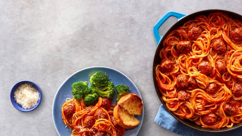 A big pot of spaghetti is shown with meatballs, to the side is a small plate of spaghetti and meatballs, brocolli and a slice of garlic bread.