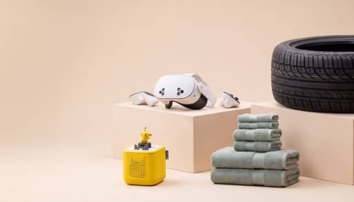 Virtual reality headset on a block beside folded green towels, a black car tire and a small yellow speaker with a toy figure.