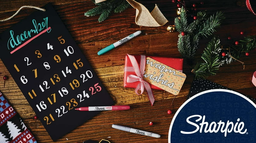 Assorted Sharpie markers sit on a wooden table with Christmas decorations and a December month calendar.