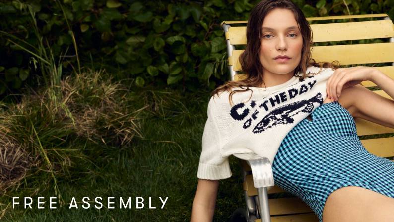 A woman in a gingham bathing suit with a short-sleeved sweater that says ‘Catch of the Day’ on it, reclining on a patio chair against greenery.