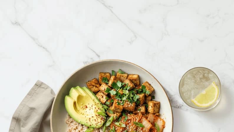 A grain bowl is shown topped with avocado & green onion. Showed next to a water cup with lemon slices.