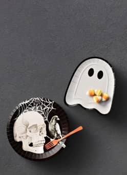 Image shows a black paper plate with a skull, cobweb and crow design and an orange plastic fork. A ghost-shaped paper plate is shown with candy.
