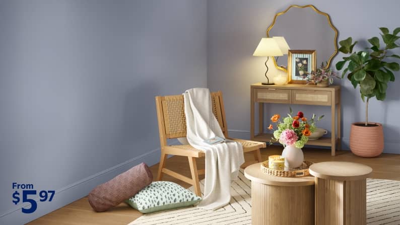 A living room with a woven chair draped with a throw blanket, wood dining table, side table, rug, ficus and pillows on the floor.