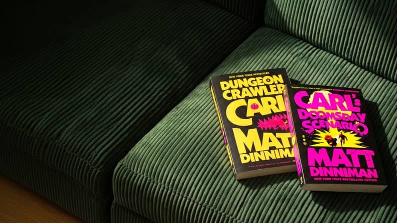Two Matt Dinniman paperback books Dungeon Crawler Carl and Carls Doomsday Scenario rest on a ribbed sofa in sunlight.