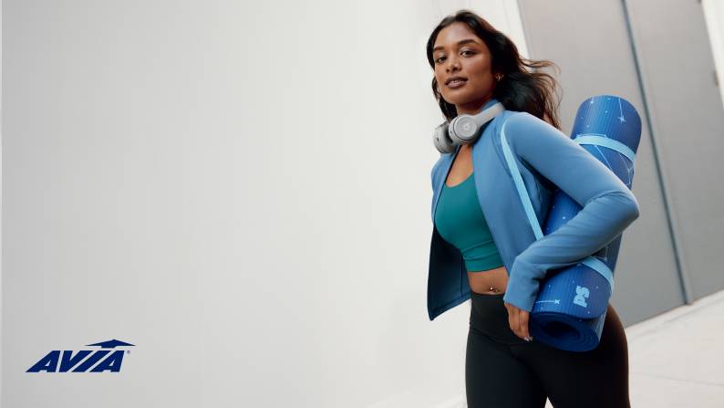 A young woman is shown wearing an Avia sports bra, blue zip-up jacket and black leggings. She is carrying a yoga mat.