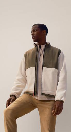 A man wearing a green and cream fleece and caramel pants is pictured on a beige backdrop.
