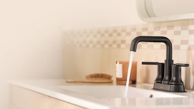 Water running from dark faucet at a white sink. A brush and candle sit beside the sink.