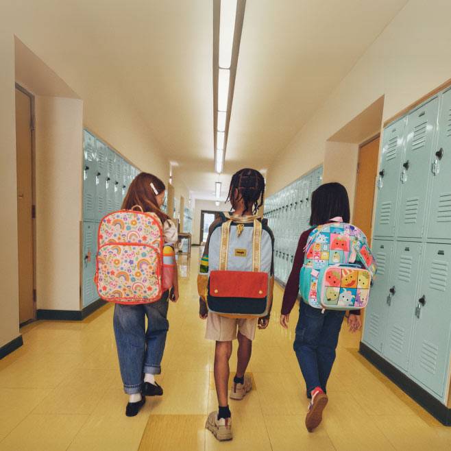 School-age kids walking down a locker hallway wearing colorful pink, blue, and checkered graphic school backpacks.