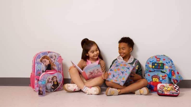 Two children sitting with notebooks and character themed backpacks, folders and school supplies.