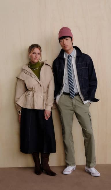 Two adults in layered fall outfits stand side by side, one in a beige jacket and skirt, the other in a denim jacket, tie and khaki pants.