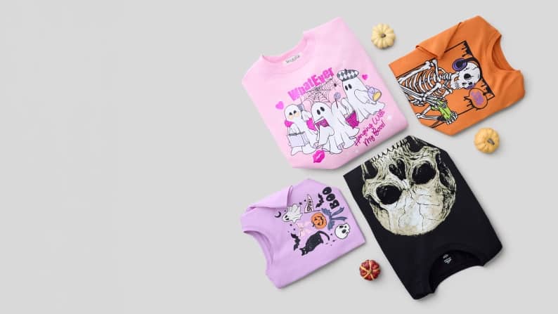 Four folded Halloween graphic T shirts with ghosts, skeletons and skull designs arranged with small decorative pumpkins.
