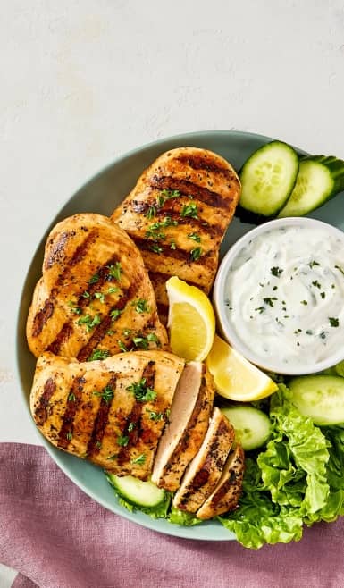 Grilled chicken breasts on a plate with lemon wedges, lettuce, cucumber slices, and a bowl of yogurt dip.
