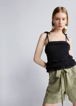 Woman with long blonde hair wearing a black ruffle trim camisole top and olive-green tie waist shorts against a light gray background.
