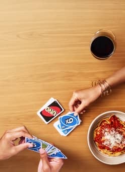 Hands playing a card game on a wooden table with a bowl of pasta nearby.