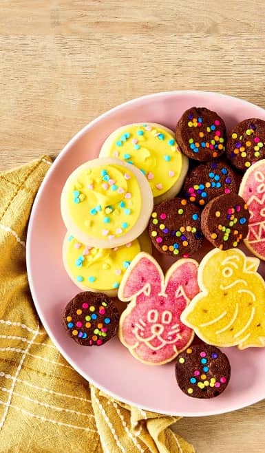 A pink plate is shown topped with yellow sugar-frosted cookies, egg-shaped cookies and mini brownie bites with sprinkles.