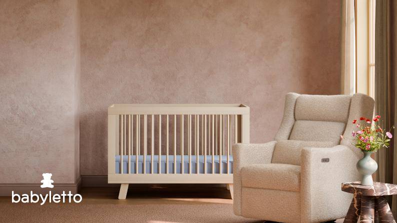Babyletto crib and upholstered nursery chair beside a small table with flowers in a minimalist nursery.