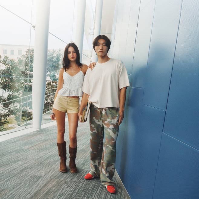 Gen Z woman and man outdoors in front of a blue wall wearing a white tank top, khaki shorts, brown boots, white tee and camo pants.