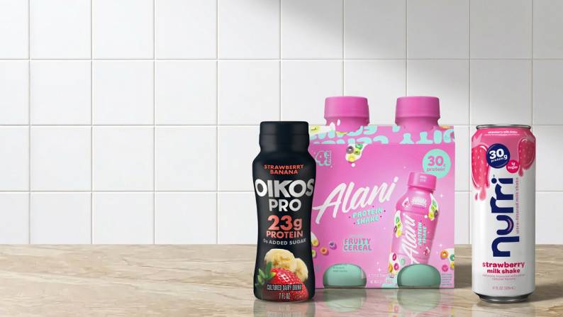 Oikos pro strawberry banana dairy drink, Alani fruity cereal protein shakes, and Nurri strawberry milkshake on a counter.