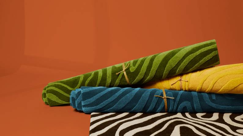 Bundled rugs are stacked and piled on top of each other. One of them is rolled out to reveal its abstract pattern.