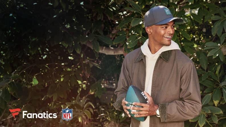 Fanatics logo and NFL logo are in the left corner. A man holding a Philadelphia Eagles football and wearing a hat is standing in front of tree.