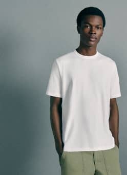 A fashion model wears a plain white short sleeve tee with light green pants against a gray background.