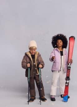 A boy and girl are shown. He wears a brown microfleece lined jacket, snowbibs, boots and a beanie. She wears a purple lined jacket, snowbibs, boots.