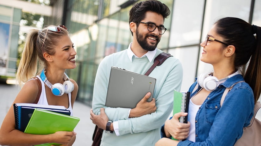 Group of three people standing outside a building holding laptops and notebooks and having a conversation.
