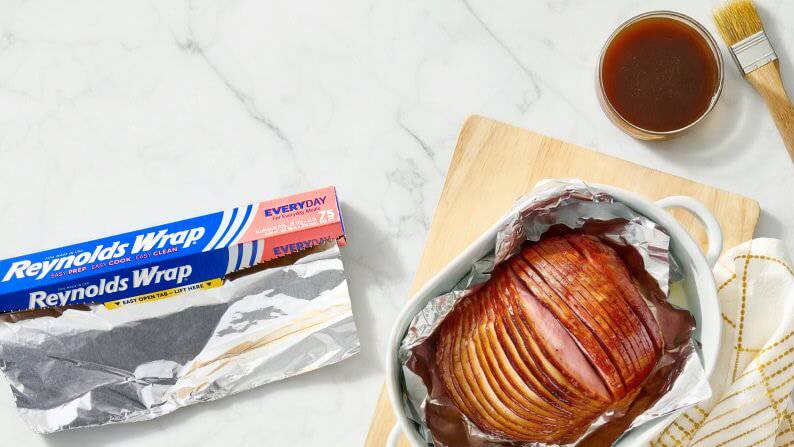 A roll of Reynolds Wrap aluminum foil is pictured next to a baked ham in a serving tray.