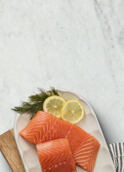 Two fillets of raw salmon with sliced lemon and fresh herbs are shown on a platter on top of a marble counter.