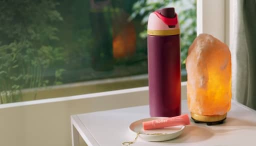 A maroon water bottle stands next to a glowing pink salt lamp and a small tube of lip balm on a white table.