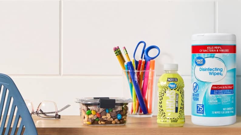 A desk with trail mix in a container, writing utensils in a cup, and Great Value products such as a bottled beverage and disinfecting wipes.