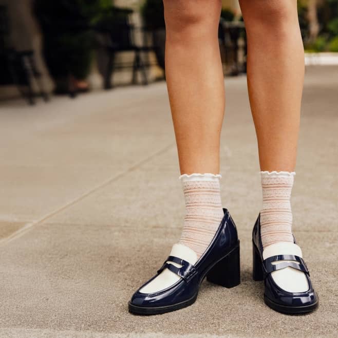 Adult wearing sheer socks and navy and white block heel loafers standing on a paved surface.