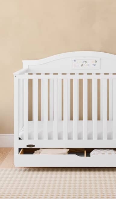White baby crib with slatted sides and open storage shelf holding folded blankets.