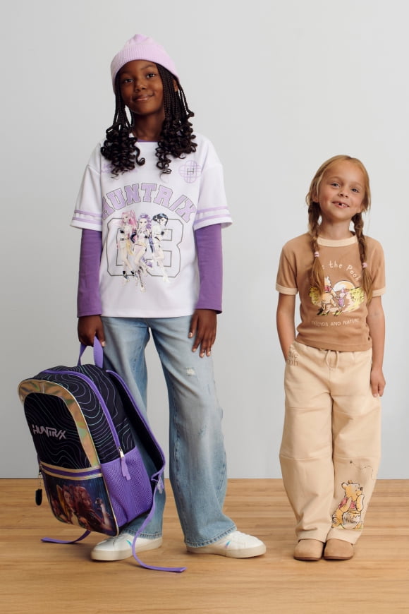 Young girls in character graphic tees, tan cargo pants, and wide-leg jeans by blue lockers; trendy back-to-school character fashion.
