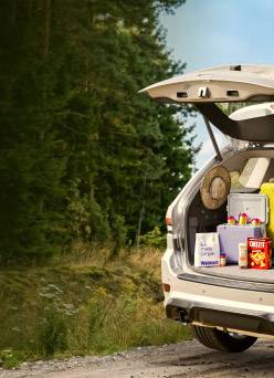 An open hatchback car filled with snacks, meds, skincare, accessories, and a suitcase is in a woodsy area.
