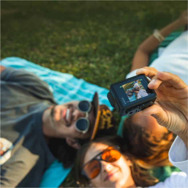 Image of three people lying on the ground outside taking a photo of themselves on a black digital camera.