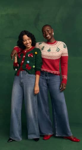 Two women pose in festive sweaters and wide-leg jeans against a dark green background.