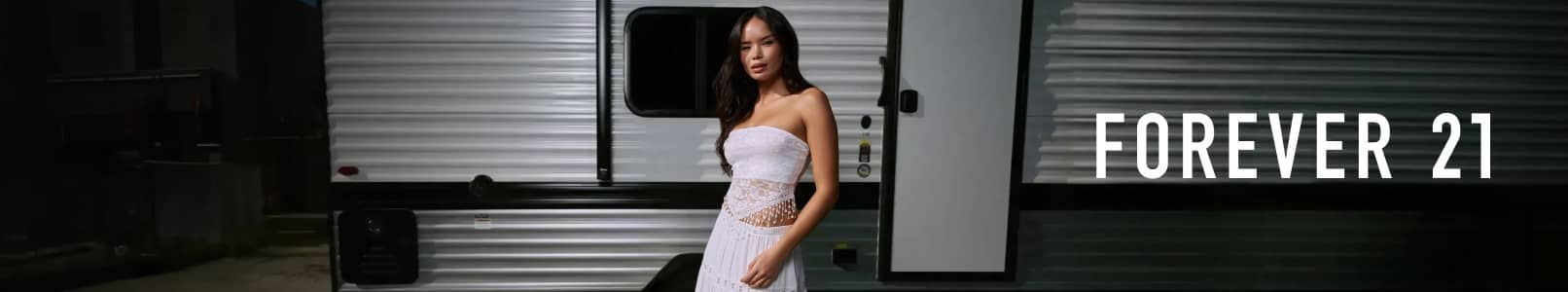 Young woman standing by an RV in a white sheer lace strapless tube top and white tiered maxi skirt. Forever 21 logo is seen.